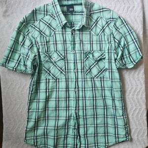 Grön rutig skjorta Jack & Jones - Kortärmad grön rutig skjorta från Jack & Jones med två bröstfickor och knappar framtill. Skjortan har klassisk krage och detaljer i svart och vitt i rutmönstret. Perfekt för en avslappnad och stilren look.