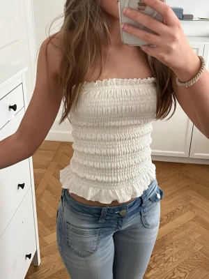 Vit bandeau tubtopp - Så söt vit tubtopp, perfekt inför sommaren🤩 Toppen är smockad och har volangkant längst ner. Materialet är stretchigt💕 Nypris: 259kr.