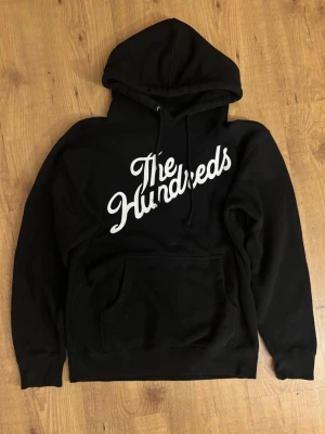 Svart hoodie The Hundreds - Svart hoodie med stor vit The Hundreds-logga på bröstet. Klassisk känguruficka framtill och justerbar huva med snören. Tillverkad i mjukt material som passar perfekt för en avslappnad streetwear-look.