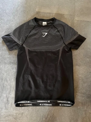 Gymshark Onyx V5 Grey Compressionshirt  - Snygg svart träningströja från Gymshark med korta ärmar och meshdetaljer över bröst och axlar. Tröjan har ett vitt Gymshark-logotyptryck på bröstet och texten 'BE A VISIONARY' längs nederkanten. Tillverkad i ett lätt och ventilerande material. Använd fåtal gånger.