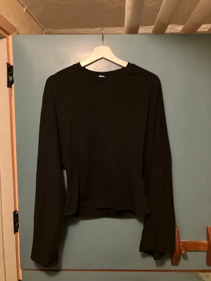 Svart långärmad topp i bomull - En svart långärmad tröja. Tröjan har rund halsringning och vida ärmar, vilket ger en chill och trendig look. Perfekt att matcha med jeans eller kjol för en enkel men snygg outfit.