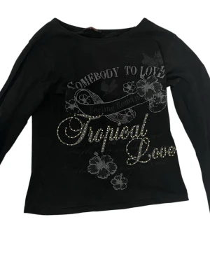Svart långärmad topp med tryck - Svart långärmad topp med texten 'Tropical Love' och blommigt tryck framtill. Dekorerad med små glittriga detaljer som ger extra edge. Rak passform och rund halsringning. Perfekt för dig som gillar statement-plagg.