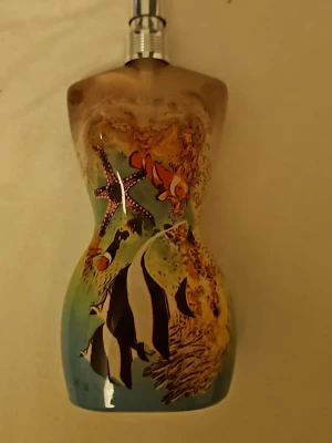 Jean Paul Gaultier parfymflaska havsmotiv - Unik parfymflaska från Jean Paul Gaultier formad som en kvinnokropp, dekorerad med färgglada havsmotiv som clownfiskar, sjöstjärnor och koraller. Flaskan har blå, gröna och beige toner och är tillverkad i glas med metallförslutning. Perfekt för dig som gillar cool design!