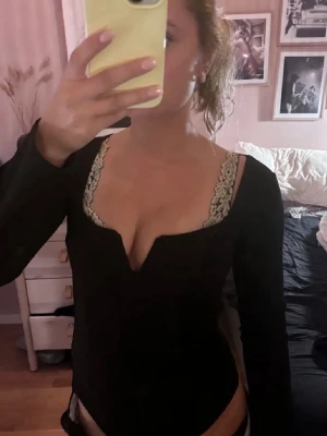 Svart långärmad body med v ringning  - Säljer en svart långärmad body med djup urringning och dekorativa spetsdetaljer i beige längs kanterna. Bodyn har en åtsittande passform och är perfekt för dig som gillar en stilren men ändå sexig look. Materialet ser ut att vara stretchigt och mjukt.