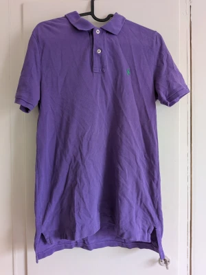 Lila pikétröja från Ralph Lauren - Säljer en klassisk lila pikétröja från Ralph Lauren med korta ärmar och krage. Tröjan har två knappar framtill och den ikoniska gröna loggan broderad på bröstet. Tillverkad i mjuk bomull som andas och har slitsar i sidorna för extra komfort.