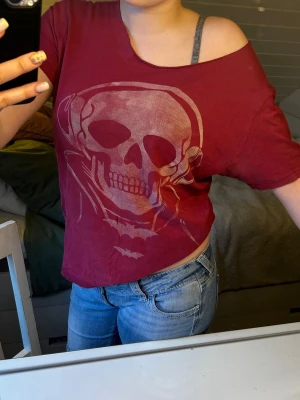 Röd offshoulder t-shirt med dödskalle - Säljer en röd t-shirt med offshoulder-design och stort dödskalletryck framtill. T-shirten har korta ärmar och en loose passform som ger en avslappnad vibe. Perfekt för dig som gillar edgy och unik stil. Den har några fläckar och hål som man kan se på bilden. 