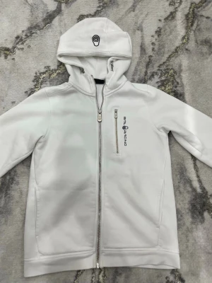 Vit hoodie från Sail Racing - Snygg vit hoodie från Sail Racing med dragkedja framtill och en vertikal bröstficka med logga. Mjuk och slät yta, huva med tryckt detalj och långa ärmar. Perfekt för en clean och sportig look.