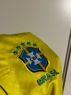 Brasilien Neymar Jr 10 matchtröja - Gul Brasilien fotbollströja med Neymar Jr och nummer 10 på ryggen. Tröjan har broderat CBF-märke med fem stjärnor och texten BRASIL på bröstet. Gröna och blå detaljer vid krage och ärmslut. Perfekt för dig som älskar fotboll och vill stötta landslaget.