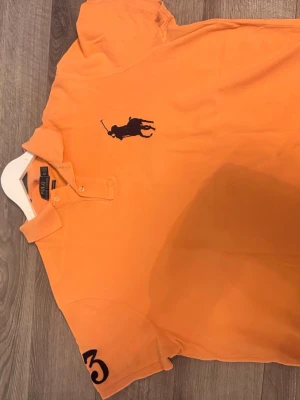 Orange pikétröja från Polo Ralph Lauren - Snygg orange pikétröja från Polo Ralph Lauren med klassisk krage och två knappar. Stor mörkblå logga på bröstet och siffran 3 på ärmen. Tröjan är i bomull och har en slim fit passform som sitter snyggt på kroppen.