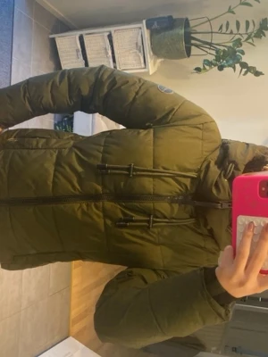 Vinter jacka  - Säljer en olivgrön pufferjacka från marikoo med huva och svarta detaljer. Jackan har dragkedja framtill, två sidofickor och justerbara snören vid huvan. Märke syns på vänster ärm. Perfekt för kalla vinterdagar och riktigt skön att bära.