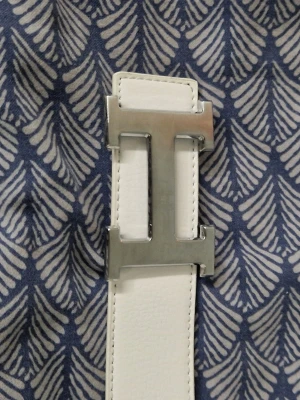 Vitt skärp med silverfärgat H-spänne - Snyggt vitt skärp med ett stort, silverfärgat H-format spänne. Skärpet har en stilren och minimalistisk design som passar till många olika outfits. Tillverkat i skinn för en exklusiv känsla.