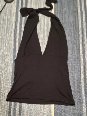 Svart halterneck linnetopp - Snygg svart linnetopp med halterneck och djup v-ringning. Toppen knyts i nacken och har en tight passform som framhäver axlarna. Perfekt för att styla med jeans eller kjol för en trendig look.