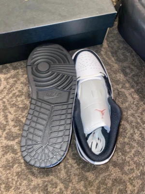 Air Jordan 1 Low svart/vit/rosa - Snygga Air Jordan 1 Low sneakers i svart, vitt och rosa. Skorna har klassisk låg siluett, vit tåbox med perforeringar, svart läder på sidorna och rosa detaljer på hälen samt logga. Tjock vit sula och svart yttersula. Perfekta för dig som gillar streetwear och ikonisk design.