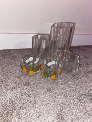 Hexagonformade glasbehållare och skålar - Säljer ett set med tre hexagonformade glasbehållare och två små glasskålar med ananasmotiv. Behållarna är transparenta och kan användas för förvaring eller dekoration. Skålarna har färgglada tryck med gula ananaser och gröna blad.