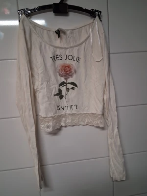 Vit långärmad topp med spetskant - Söt vit långärmad topp med tryck av en rosa ros och texten 'TRÈS JOLIE ISN'T IT?'. Toppen har spetskant nedtill och en avslappnad passform. Perfekt för dig som gillar romantiska detaljer och blommigt motiv.