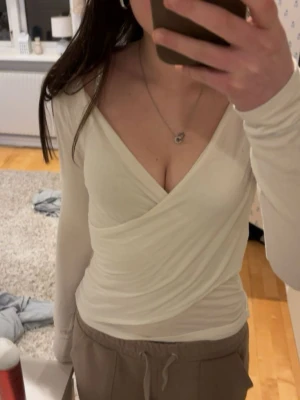 Vit omlottopp med v-ringning - Säljer en stilren vit omlottopp med djup v-ringning och långa ärmar. Toppen har en figurnära passform och är tillverkad i mjukt, stretchigt material som känns skönt mot huden. Perfekt att matcha med både jeans och kjol. Har en liten fläck(se bild) men som knappast syns 