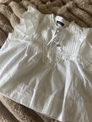 Vit poplinblus från Gina Tricot - Supersöt vit poplinblus från Gina Tricot med volangdetaljer och knytning vid halsen. Blusen har fina veck framtill och korta ärmar med volanger som ger en romantisk känsla. Perfekt att styla med jeans eller kjol.