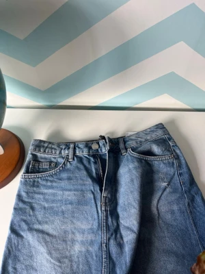 Blå jeanskjol med klassisk passform - Säljer en blå jeanskjol med klassisk femficksdesign och knappgylf. Kjolen har en rak modell och är tillverkad i slitstark denim, perfekt för en avslappnad och trendig look.