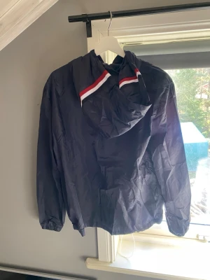 Svart vindjacka från Moncler - Snygg svart vindjacka från Moncler med huva. Jackan har en cool detalj med röd och vit rand på huvan. Tillverkad i lätt och vindtätt material, perfekt för blåsiga dagar. Enkel och stilren design som passar till det mesta.