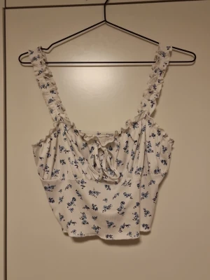 Blommig vit croppad linnetopp - Supersöt croppad linnetopp i vitt med blått blommönster och volangdetaljer på axelbanden. Toppen har en söt knytning framtill och är perfekt för varma dagar. Materialet känns mjukt och lätt, troligen bomull eller polyester.