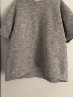 Grå stickad t-shirt i ullmix - Mysig grå stickad t-shirt med korta ärmar och rund hals. Oversized passform och mjukt material, perfekt för lager på lager. Enkel och stilren design som passar till det mesta.