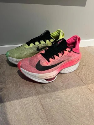 Nike ZoomX Vaporfly Next% 2 sneakers - Nike ZoomX Vaporfly Next% 2 sneakers med en sko i neonrosa och en i neongrönt. Båda har svart swoosh, svarta skosnören och vit, tjock sula med ZoomX-dämpning. Ovandelen är i mesh för bra ventilation och lätt känsla.