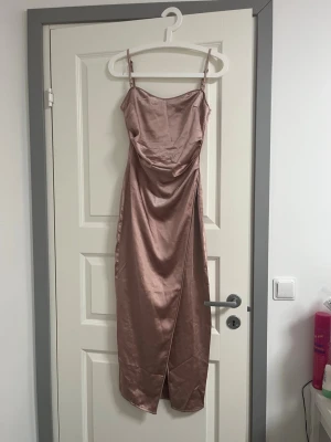 Champagnefärgad långklänning i satin - Säljer en elegant långklänning i champagnefärgad satin med smala axelband och draperad front. Klänningen har en slits framtill och en figurnära passform som ger en snygg siluett. Perfekt för dig som vill ha en stilren och trendig look.