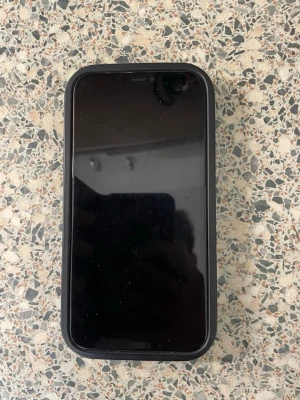 Svart iPhone 11 med svart skal - Säljer en svart iPhone med ett svart skyddsskal. Telefonen har rundade hörn och en stor touchskärm. Klassisk Apple-design med stilren look och glas på framsidan. Perfekt för dig som vill ha en enkel och snygg mobil.