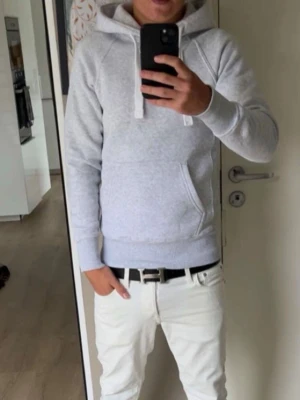 Ljusgrå hoodie med huva - Säljer en stilren ljusgrå hoodie med huva och vita dragsnören. Hoodien har en klassisk känguruficka framtill och ribbade muddar vid ärmslut och nederkant. Perfekt för en avslappnad och trendig look.