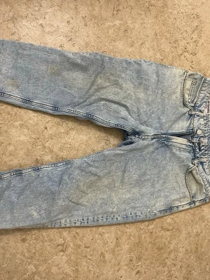 Ljusblå mom jeans i denim - Säljer ett par ljusblå mom jeans i klassisk denim med hög midja och raka ben. Jeansen har fem fickor och stängs med dragkedja och knapp. Perfekta för en avslappnad och trendig look.