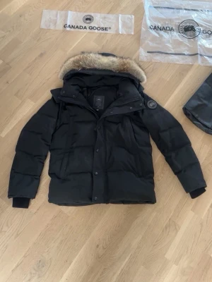 Svart dunjacka från Canada Goose - Snygg svart dunjacka från Canada Goose med avtagbar huva och pälsdetalj. Jackan har klassisk patch på ärmen och stora fickor 