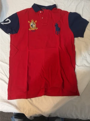 Röd pikétröja från Ralph Lauren - Klassisk röd pikétröja från Ralph Lauren med mörkblå krage och ärmar. Stort blått logobroderi på bröstet och broderad patch med siffran 2 på ena ärmen. Tillverkad i bomull och har knappar vid halsen.