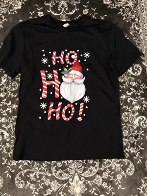 Svart jul-tshirt med tomte och HO HO HO - Svart t-shirt med tryck av tomte och texten HO HO HO i rödvitrandigt godisstil. T-shirten har korta ärmar och rund hals. Perfekt för dig som vill maxa julkänslan med en rolig och färgglad design.