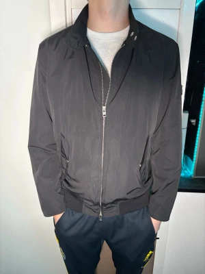 Svart J.Lindeberg jacka  - Snygg svart jacka från J.Lindeberg med dragkedja framtill, två snedställda fickor med tryckknappar och ribbade muddar vid ärmslut och nederkant. Jackan har en diskret logga på ärmen och ett tunt, glansigt innerfoder.