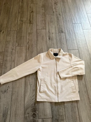 Beige overshirt med dragkedja - Stilren och enkel beige overshirt med dragkedja framtill och två fickor. Jackan har klassisk krage och raka, långa ärmar. Perfekt för lager på lager och en clean look.