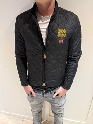 Svart quiltad jacka från Morris - Snygg svart quiltad jacka från Morris med gul broderad logga och brittisk flagga på bröstet. Jackan har dragkedja framtill, ståkrage och en slim passform. Perfekt för dig som gillar stilrena och klassiska plagg. Jackan passar dig som är 165-180 lång✅skriv vid minsta fundering😃