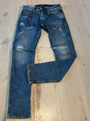 Blå slitna jeans från Replay - Säljer ett par blå jeans från Replay med slitningar och hål för en trendig look. Jeansen har fem fickor, knappgylf och raka ben. Materialet är klassisk denim i bomull med snygg tvätt och detaljerade slitningar på både fram- och baksida. Midja 76cm och längd 103 cm. Skriv om du har minsta lilla fundering😃