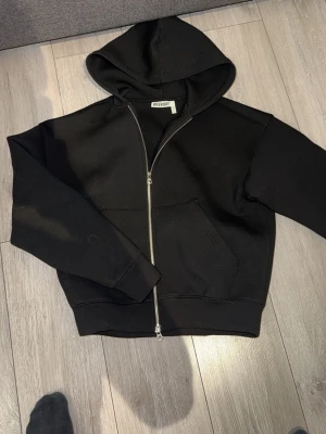 Svart hoodie med dragkedja från Weekday - Svart hoodie från Weekday i storlek XS. Modellen har dragkedja framtill, huva och känguruficka. Perfekt för en avslappnad och stilren look. Tillverkad i mjukt material som känns skönt mot huden.