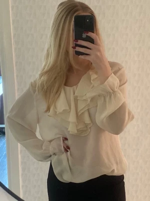 Krämvit blus med volangdetaljer - Snygg krämvit blus med långa ärmar och stora volangdetaljer framtill. Blusen har en lös passform och rund halsringning. Tillverkad i ett lätt, mjukt tyg. Står ingen storlek i tröjan men skulle säga den sitter som M. Köpt på Stirling Cooper i London.