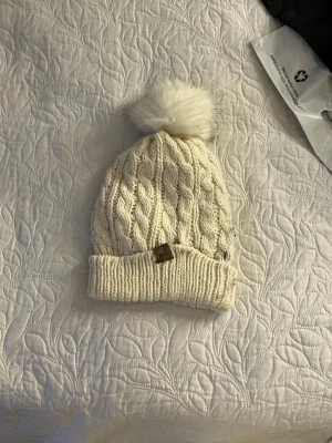 Vit stickad mössa med tofs - Säljer en krämvit stickad mössa med flätmönster och stor fluffig tofs upptill. Mössan har uppvikt kant och en liten brun logolapp framtill. Perfekt för kalla dagar och riktigt mysig stil.