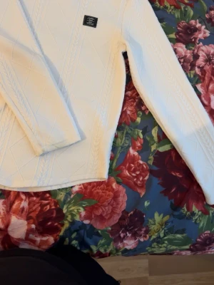 Vit stickad tröja med strukturmönster - Säljer en vit stickad tröja med snyggt strukturmönster i form av rutor och flätor. Tröjan har rund halsringning, långa ärmar och en liten svart patch på bröstet. Perfekt för dig som gillar stilrena och klassiska plagg.