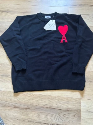 Svart stickad tröja från Ami - Snygg svart stickad tröja från Ami med röd hjärtlogga och bokstaven A på bröstet. Tröjan har rund halsringning, ribbade muddar och nederkant. Perfekt för dig som gillar stilrena plagg med en twist.