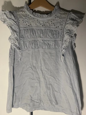 Ljusblå blus med spetsdetaljer - Supersöt ljusblå blus med broderade spetsdetaljer och volang vid axlarna. Andvänd kanske en gång och är som ny! Blusen har hög krage och fina hålbroderier upptill samt rynkade partier framtill. Perfekt för dig som gillar romantisk och feminin stil. Storlek S men känns mer som XS