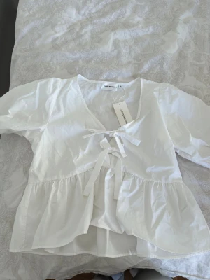 Vit blus med knyt från Carin Wester - Söt vit blus från Carin Wester med v-ringning och knytband framtill. Blusen har volang nedtill och puffiga korta ärmar.