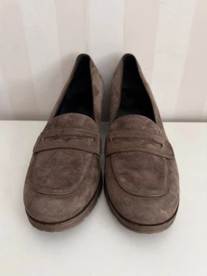 Ara Bruna mockaloafers med klack 39 - Säljer ett par snygga loafers i brun mocka med klassisk pennyloafers-design och låg klack. Skorna har rundad tå och diskreta sömmar för en stilren look. Perfekta för dig som vill ha en tidlös och trendig sko till vardagslooken.