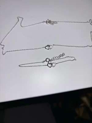 Silverfärgat smyckeset med hjärtan - Säljer ett tunt silverfärgat halsband med två sammanlänkade hjärtan i mitten. Ena hjärtat är dekorerat med små glittrande stenar för extra detalj. Kedjan är finlänkad och har ett klassiskt lås. Perfekt för dig som gillar stilrena och söta smycken.