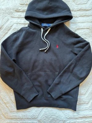 Svart hoodie från Polo Ralph Lauren - Klassisk svart hoodie från Polo Ralph Lauren med vit dragsko i huvan och röd broderad logga på bröstet. Hoodien har känguruficka framtill och ribbade muddar vid ärmar och nederkant. Tillverkad i mjukt bomullsmaterial för skön känsla.