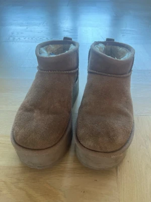 Bruna platåtofflor med foder - Mysiga bruna tofflor med hög platåsula och mjukt vitt foder på insidan. Utsidan är i mockaimitation och de har en enkel, stilren design med rund tå och låg ankelhöjd. Perfekta för att hålla fötterna varma hemma eller ute.