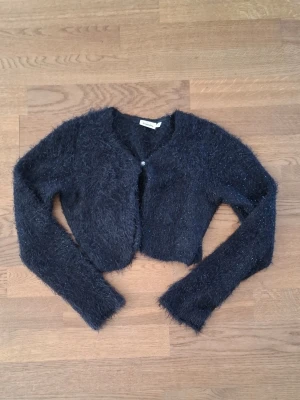 Svart fluffig cropped kofta - Mysig svart fluffig kofta från Kappahl med cropped passform och långa ärmar. Koftan har en knapp framtill och är tillverkad i ett mjukt, lurvigt material. Har storlek 158/164, men passar mig med storlek 36 och S utmärkt. 