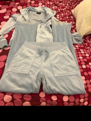 Ljusblå velourset från Juicy Couture - Säljer ett ikoniskt ljusblått velourset från Juicy Couture. Setet består av en hoodie med dragkedja och stora fickor samt matchande byxor med snörning i midjan och fickor fram. Perfekt för chill dagar hemma eller när du vill vara extra comfy.